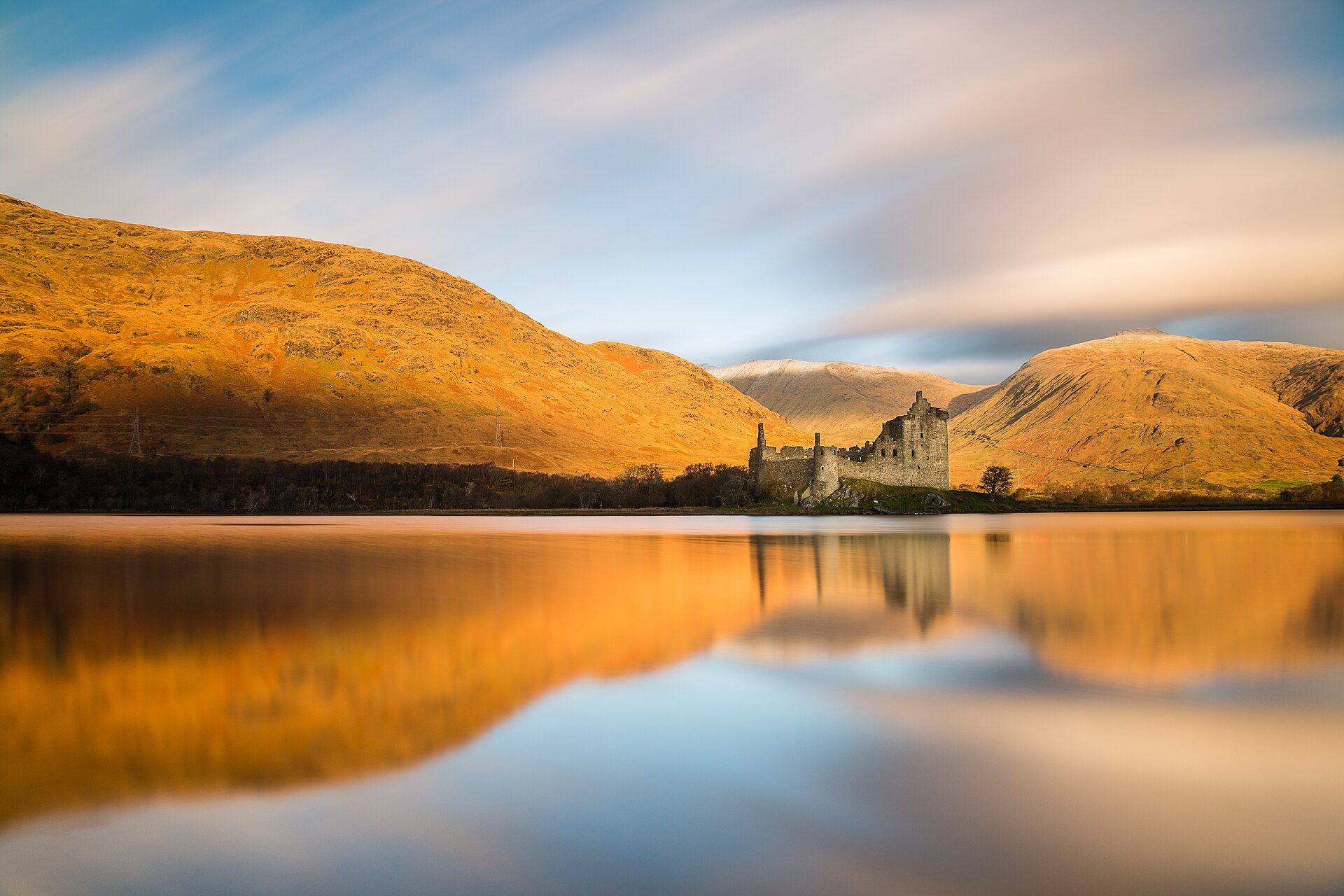 Kilchurn Castle reflected in the still water of Loch Awe at sunrise, hills behind.